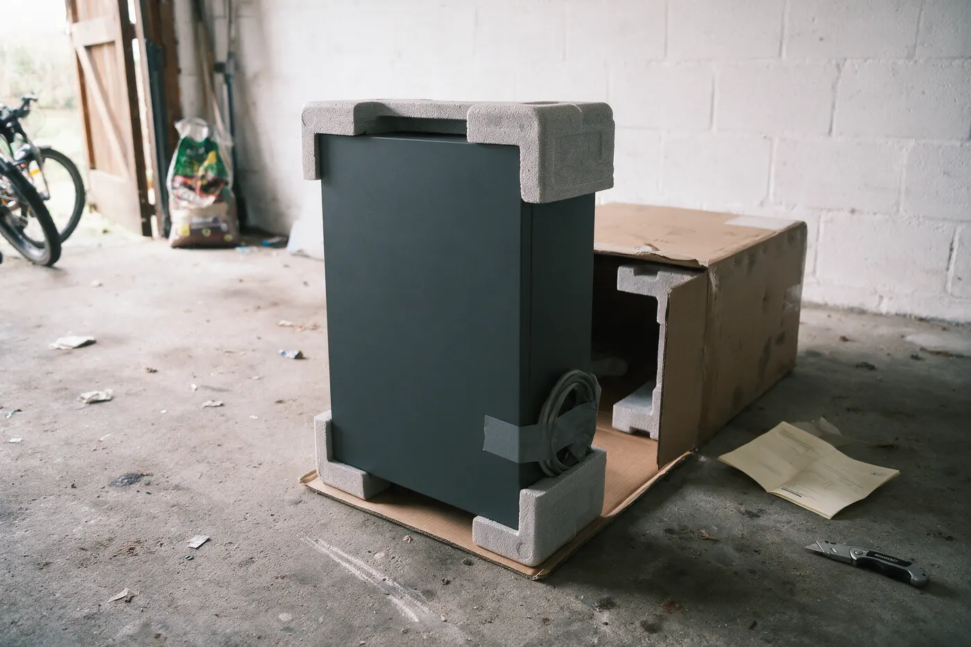 Thuisbatterij zelf installeren: een onbedrukte batterijkast half uit de kartonnen doos op een garagevloer